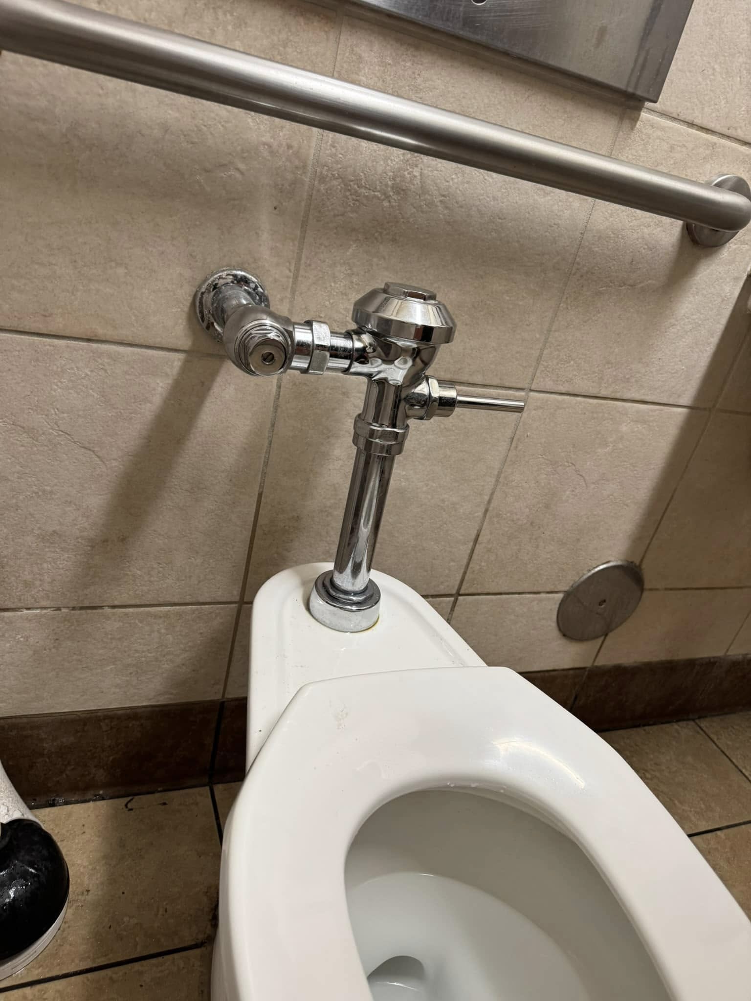 Close-up of a toilet with a flush valve and a metal grab bar mounted on the beige tiled wall in a public restroom.
