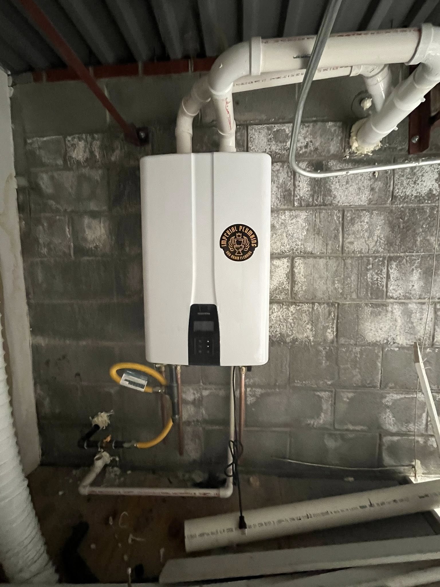A white tankless water heater is mounted on a cinder block wall, with connected pipes and a sticker reading Bison Pride on the unit.