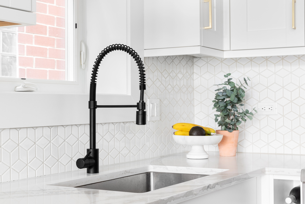 Modern kitchen sink with a matte black spring pull-down faucet, white hexagonal tile backsplash, marble countertop, and a bowl of fruit next to a potted plant—ideal for those interested in expert toilet sink and faucet installation and repair.
