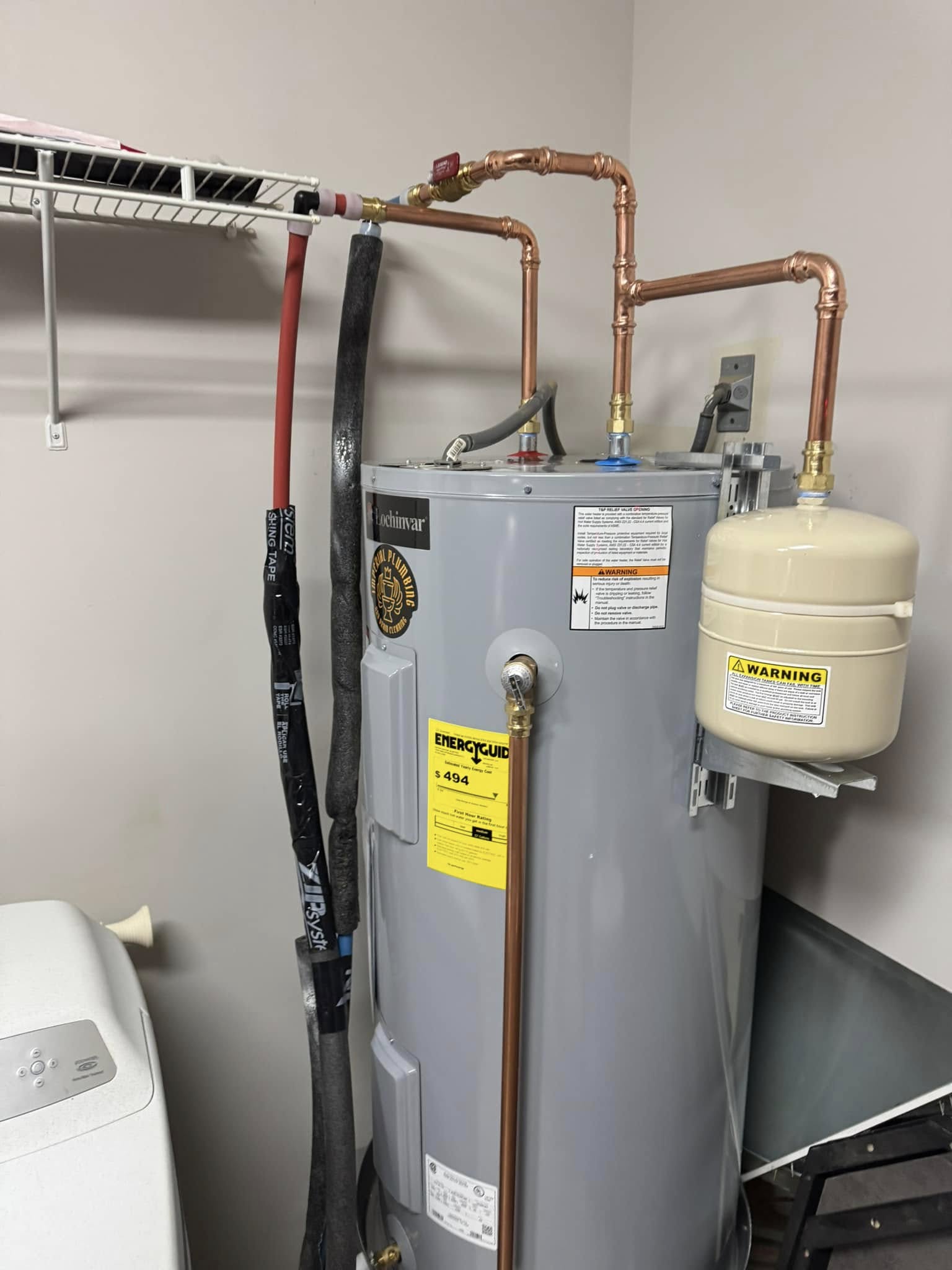 Gray water heater with attached pipes and expansion tank, positioned next to a washing machine and beneath a wire shelf in a utility room.